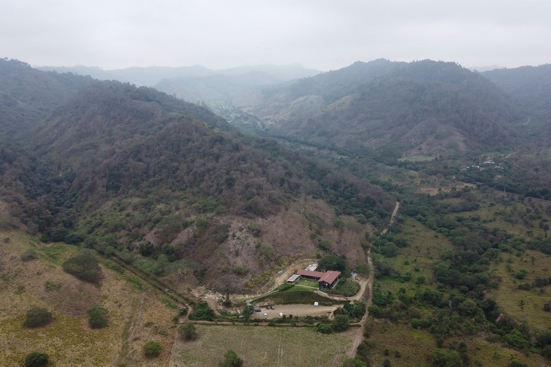 Aerial view of a single-story structure with a pond nestled in a mountainous valley under misty skies