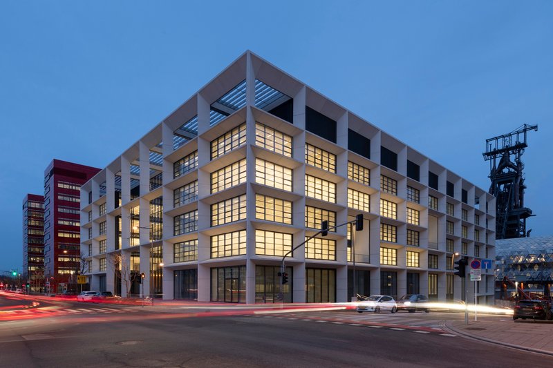 Street corner view at dusk showing the gridded facade with an industrial tower visible beyond