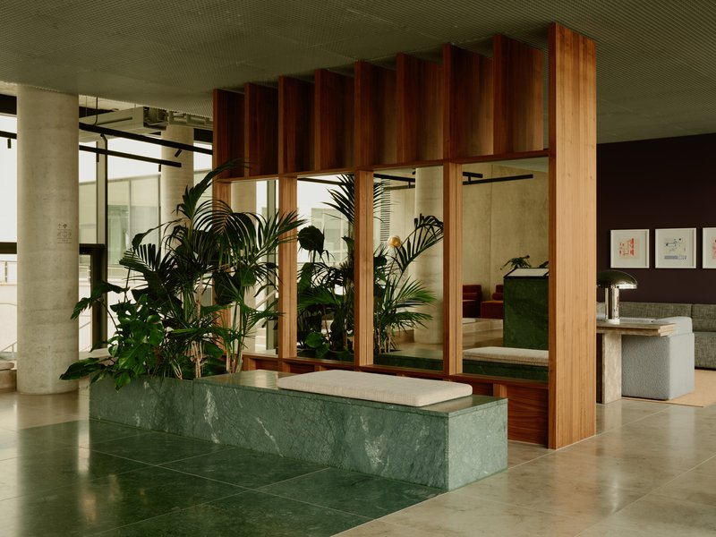 Green marble reception counter beneath a suspended timber slat installation with indoor palms