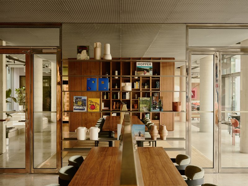 Walnut dining table facing a timber shelving wall with glass doors opening to adjacent office spaces