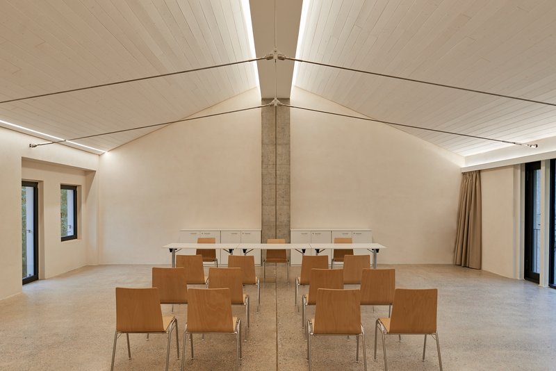 Meeting room with vaulted timber ceiling, central concrete column and rows of plywood chairs on terrazzo floor