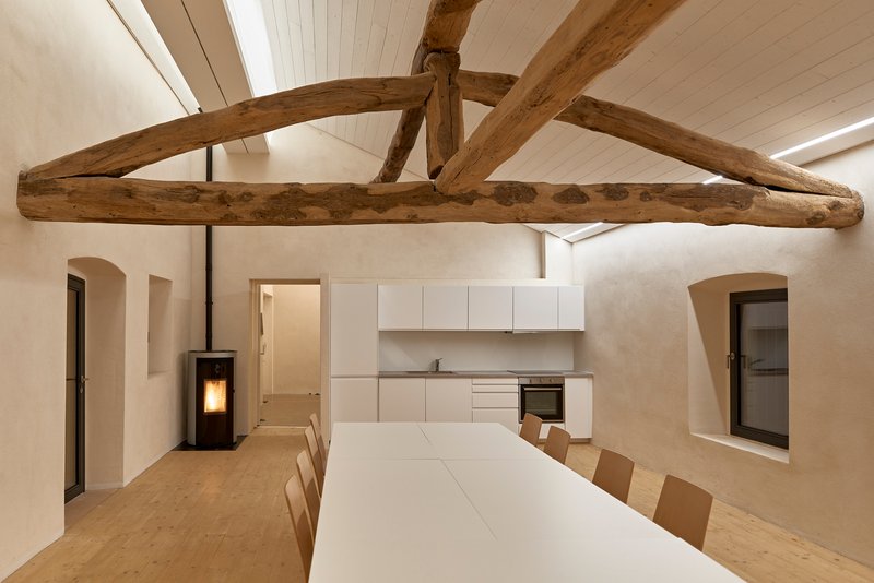 Dining area with exposed timber roof truss above white table and kitchen cabinetry along rear wall