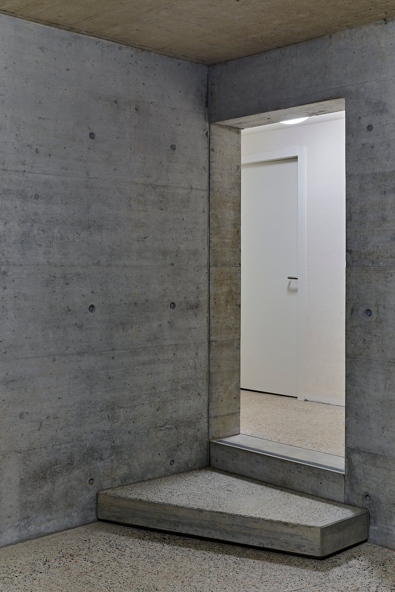 Board-formed concrete entryway with stepped platform and white door opening in dim interior light