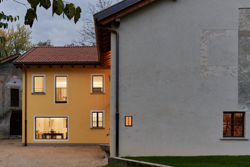 Courtyard view of illuminated yellow house and grey rendered addition at dusk