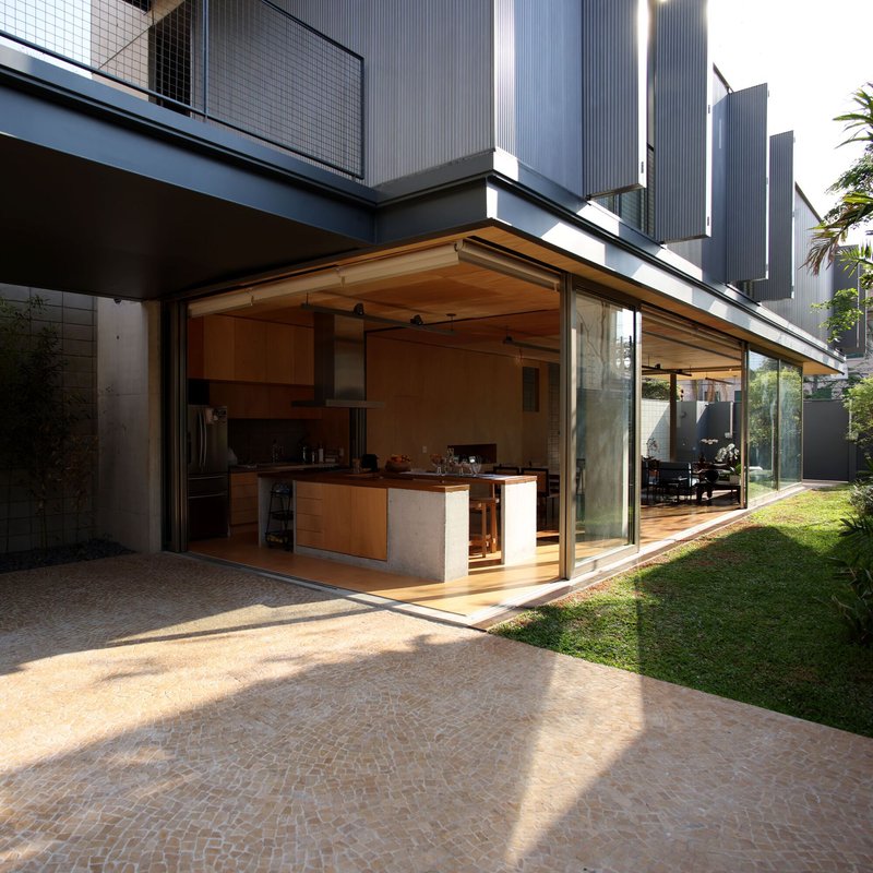 Open kitchen beneath timber-lined canopy extending from glass walls to brick-paved terrace at dusk