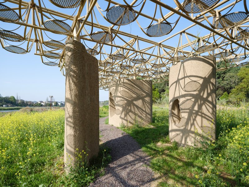 Bamboo lattice canopy supported by cylindrical columns casting dappled shadows on a gravel path through wildflowers