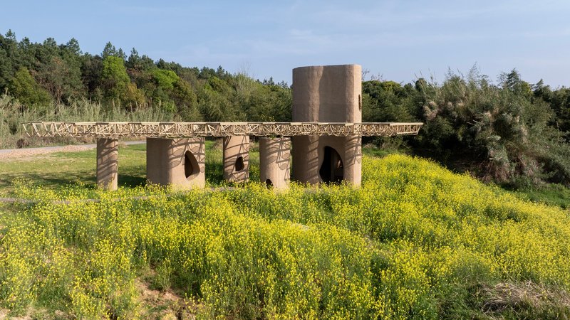 Cylindrical earthen columns with arched openings beneath a latticed canopy surrounded by blooming yellow wildflowers