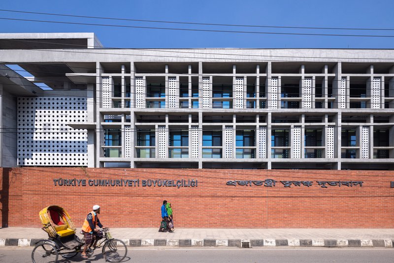Street view of the perforated white facade with horizontal concrete frame above red brick boundary wall