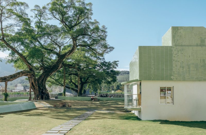 White plastered pavilion with green ceramic tile roof beside a stepping-stone path and old fig tree