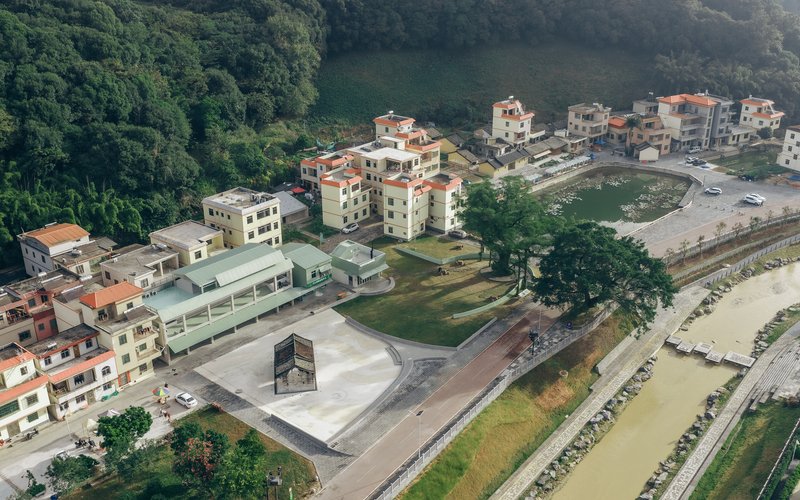 Aerial view of the village center with the elongated green-roofed building and concrete plaza beside the river