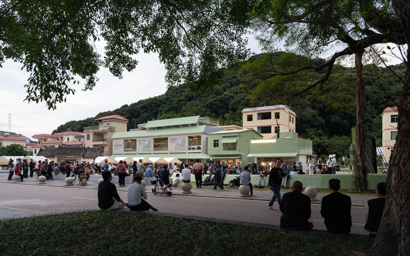 Evening view of the pavilion with open glass facade and crowd gathering on the plaza