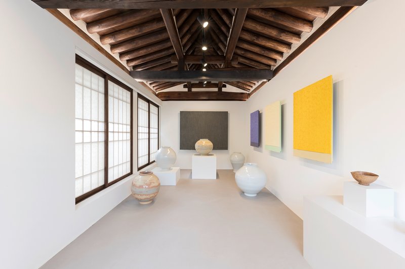 Gallery room with white ceramic vessels on pedestals and colorful paintings beneath timber truss ceiling