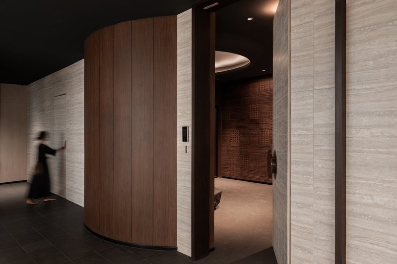 Curved wood-clad volume with elevator entrance surrounded by textured stone walls and a passing figure