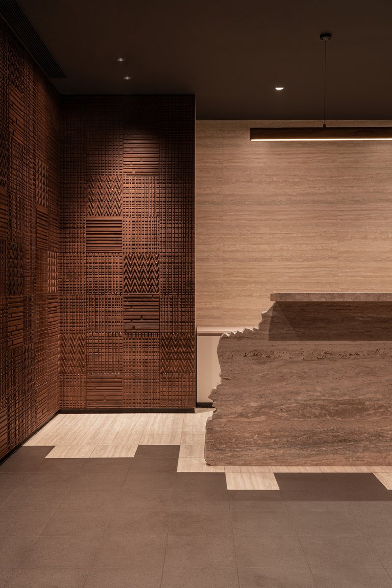 Textured copper tile wall adjacent to travertine reception desk with horizontal pendant light fixture