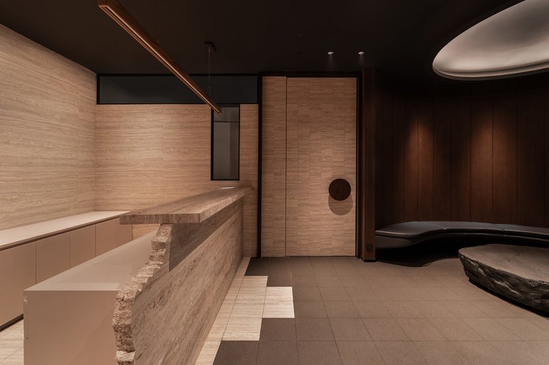 Layered travertine reception desk with rough-hewn edges beneath horizontal timber wall panels and dark ceiling