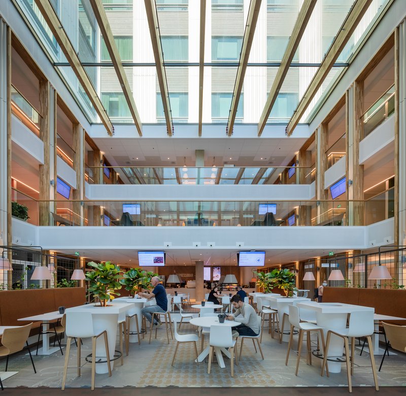 Multi-level atrium cafe with angled glass roof and people seated at white tables