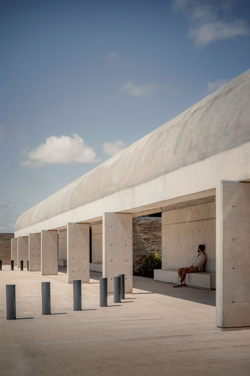 Concrete colonnade with square columns beneath a vaulted roof, with a seated figure in afternoon light