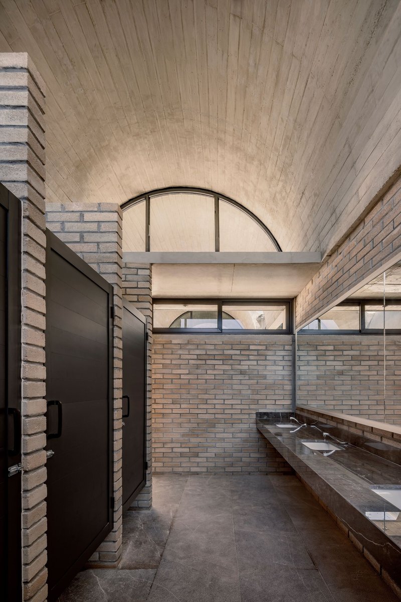 Restroom interior with tan brick walls, concrete vanity with dual sinks under vaulted ceiling