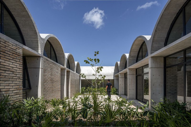 Planted courtyard between vaulted concrete volumes with brick walls and a figure standing beneath open sky