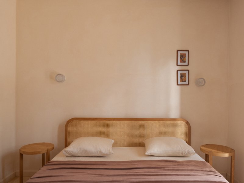 Bedroom wall with cane headboard and two framed artworks above cylindrical timber side tables