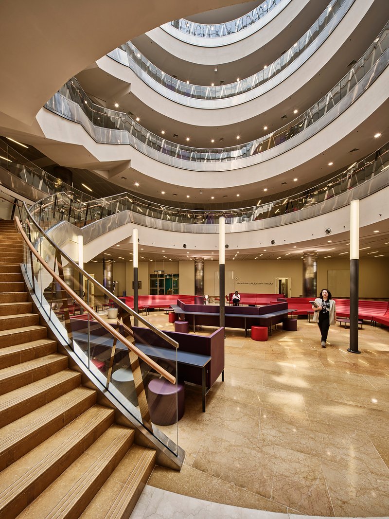 Multi-story atrium with curved balconies, timber and glass railings, and a person crossing the ground floor