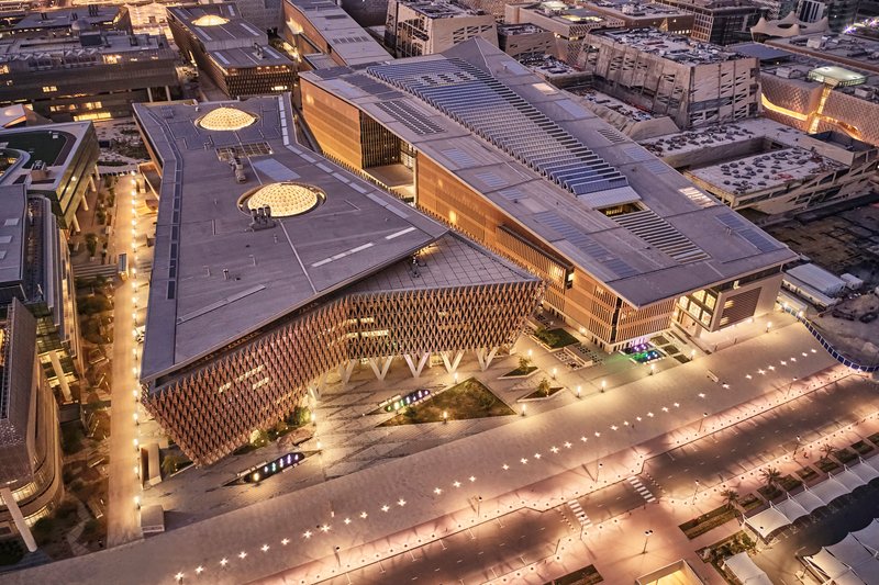 Aerial view at dusk of the complex showing illuminated plaza, lattice facade, and surrounding parking structures