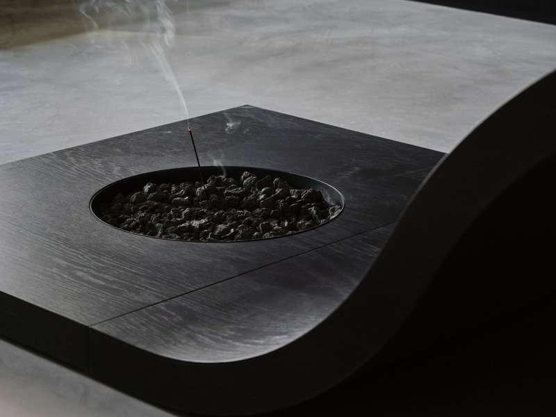 Incense stick burning above black stones in a circular recess on the dark timber surface