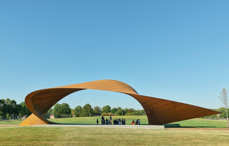 Twisted timber canopy spanning a lawn with visitors gathered beneath under clear blue sky