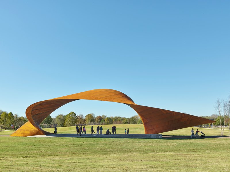 Twisted weathering steel canopy spanning a lawn with visitors gathered beneath on a clear afternoon