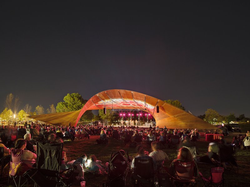Nighttime concert beneath the illuminated shell roof with crowd gathered on the grass facing the stage