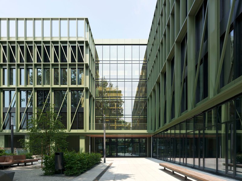 Glass skybridge connecting two wings with diagonal bracing over a courtyard entry and wooden benches