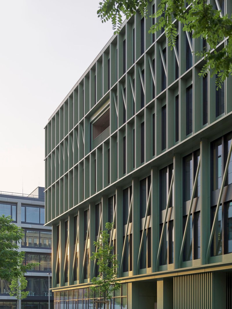 Corner facade with green vertical fins and diagonal bracing framed by street trees under overcast sky