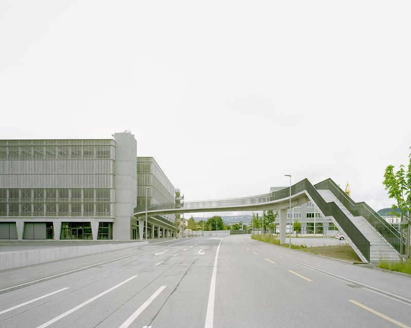 Pedestrian bridge crossing the roadway between two glass and concrete volumes under an overcast sky