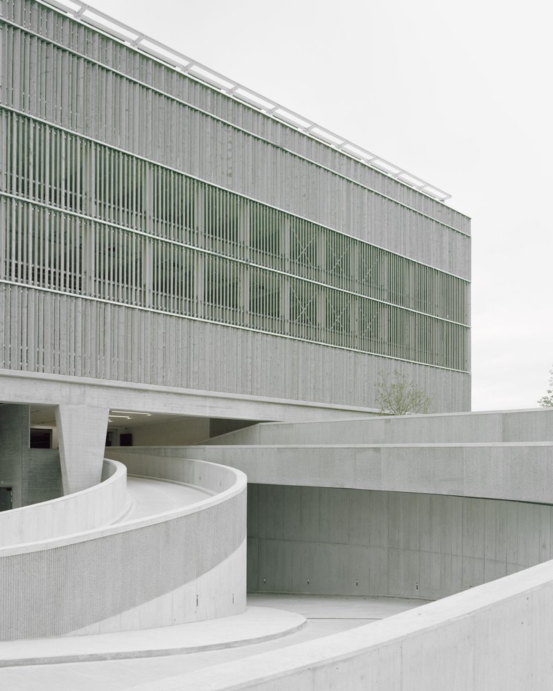 Curved concrete ramp walls beneath the louvered facade with a small tree emerging from the courtyard