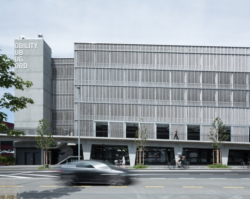 Facade with vertical louvers and canted concrete columns at street level as a car passes in the foreground