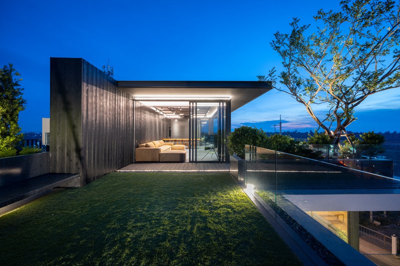 Rooftop pavilion with glass walls and timber cladding at dusk with a lawn and tree