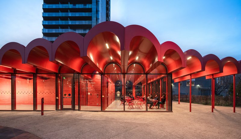 Glass facade with dark steel frames beneath the scalloped red metal roof at dusk