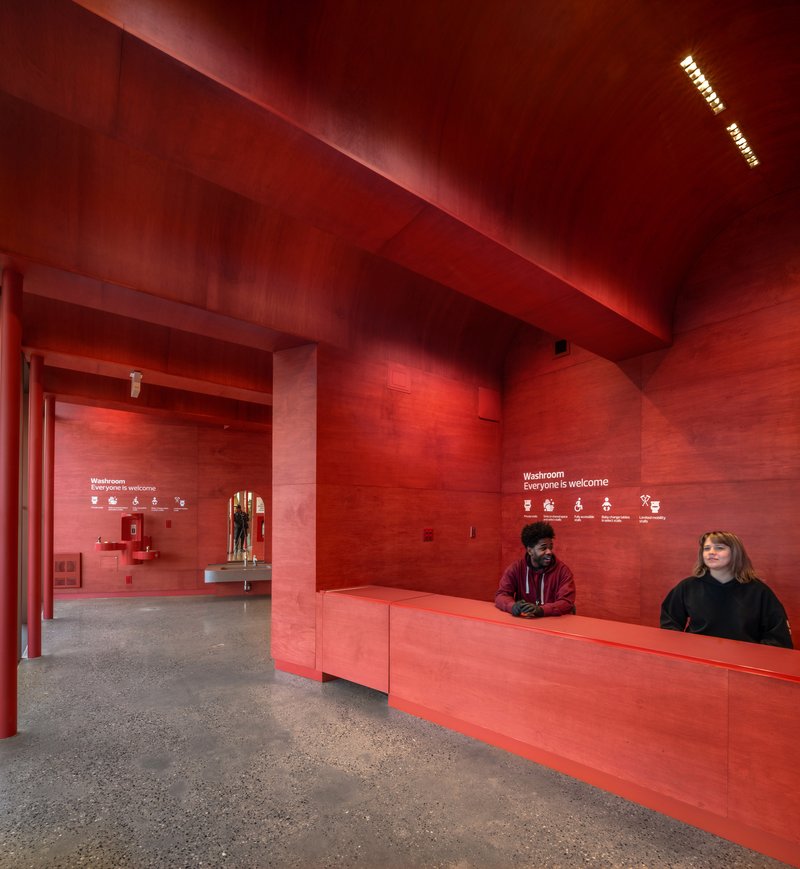 Red plywood entrance lobby with reception desk and two staff members beneath a vaulted ceiling