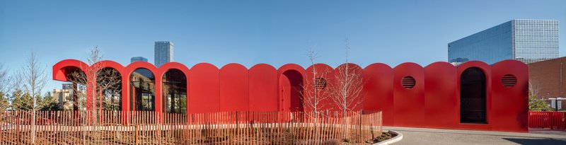 Long red facade with repeating arched profiles and timber fencing along the entry plaza