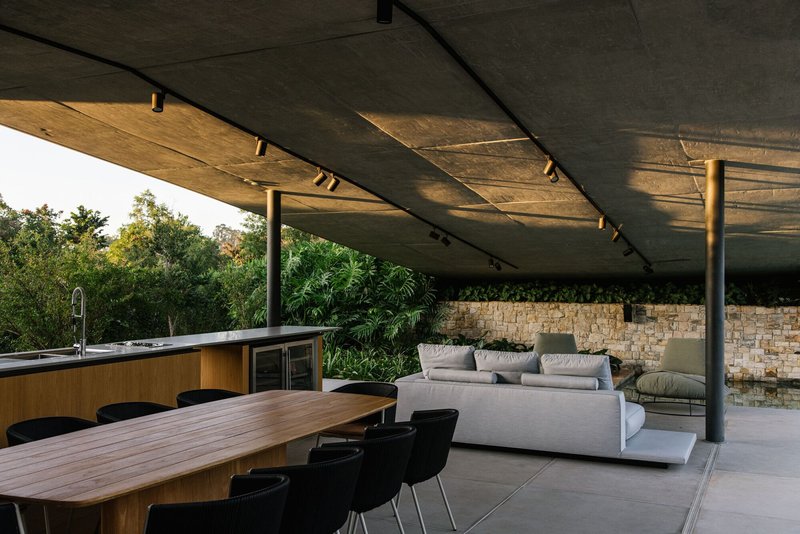 Covered terrace with concrete ceiling, steel columns, dining table and stone accent wall at sunset