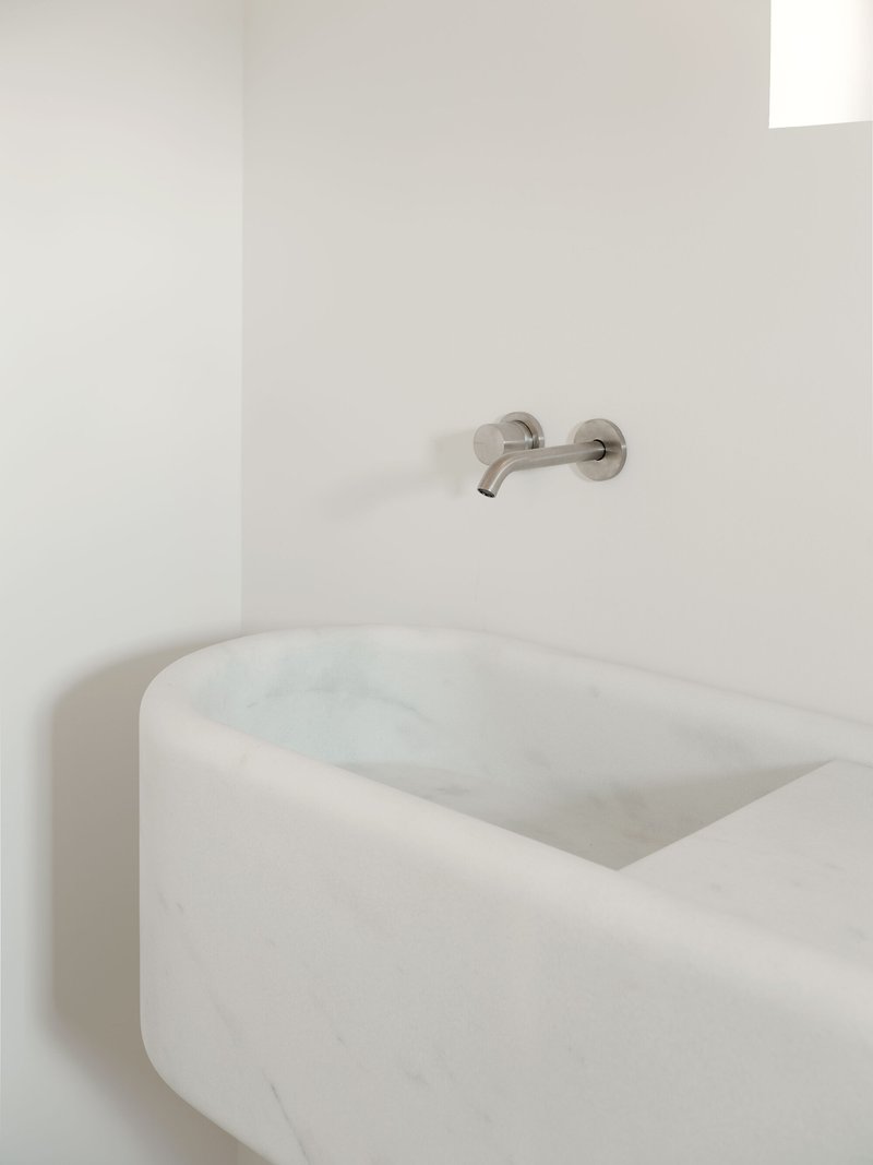 Carved marble basin with rounded edges below a wall-mounted stainless steel faucet