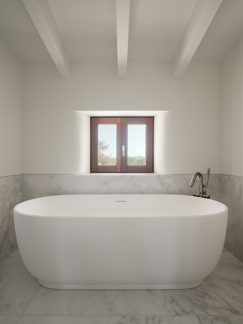 Freestanding oval bathtub beneath a deep-set timber-framed window with exposed ceiling beams