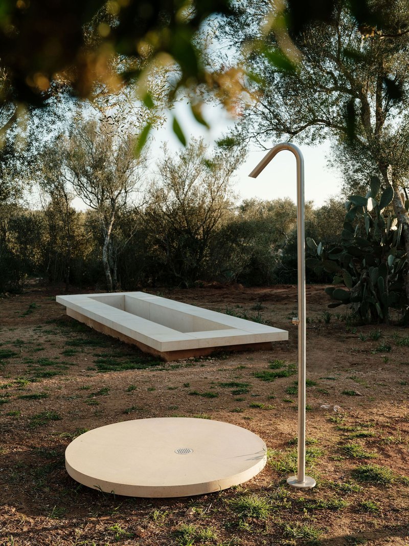 Outdoor shower with circular stone platform and curved metal fixture beside rectangular pool among olive trees