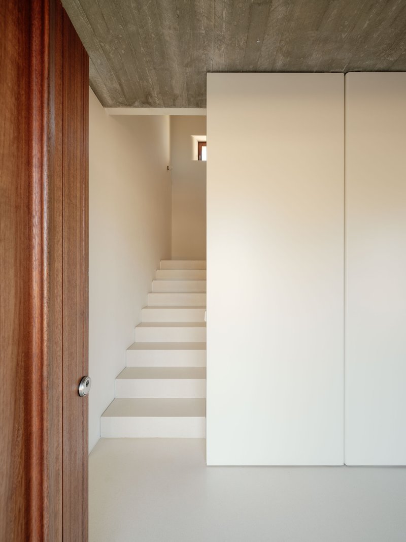 Plastered staircase rising between white walls beneath board-formed concrete ceiling