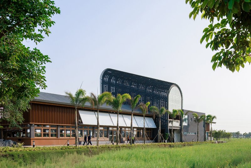 Arched volume with perforated dark cladding rising above timber pavilion and green field