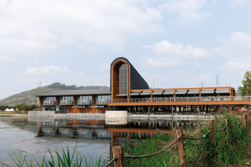 Waterside facade with barrel-vaulted volume and timber-clad wings reflecting in shallow wetland under cloudy skies