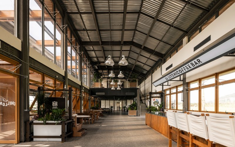 Double-height interior concourse with corrugated metal ceiling, steel columns, mezzanine and pendant lighting over cafe spaces