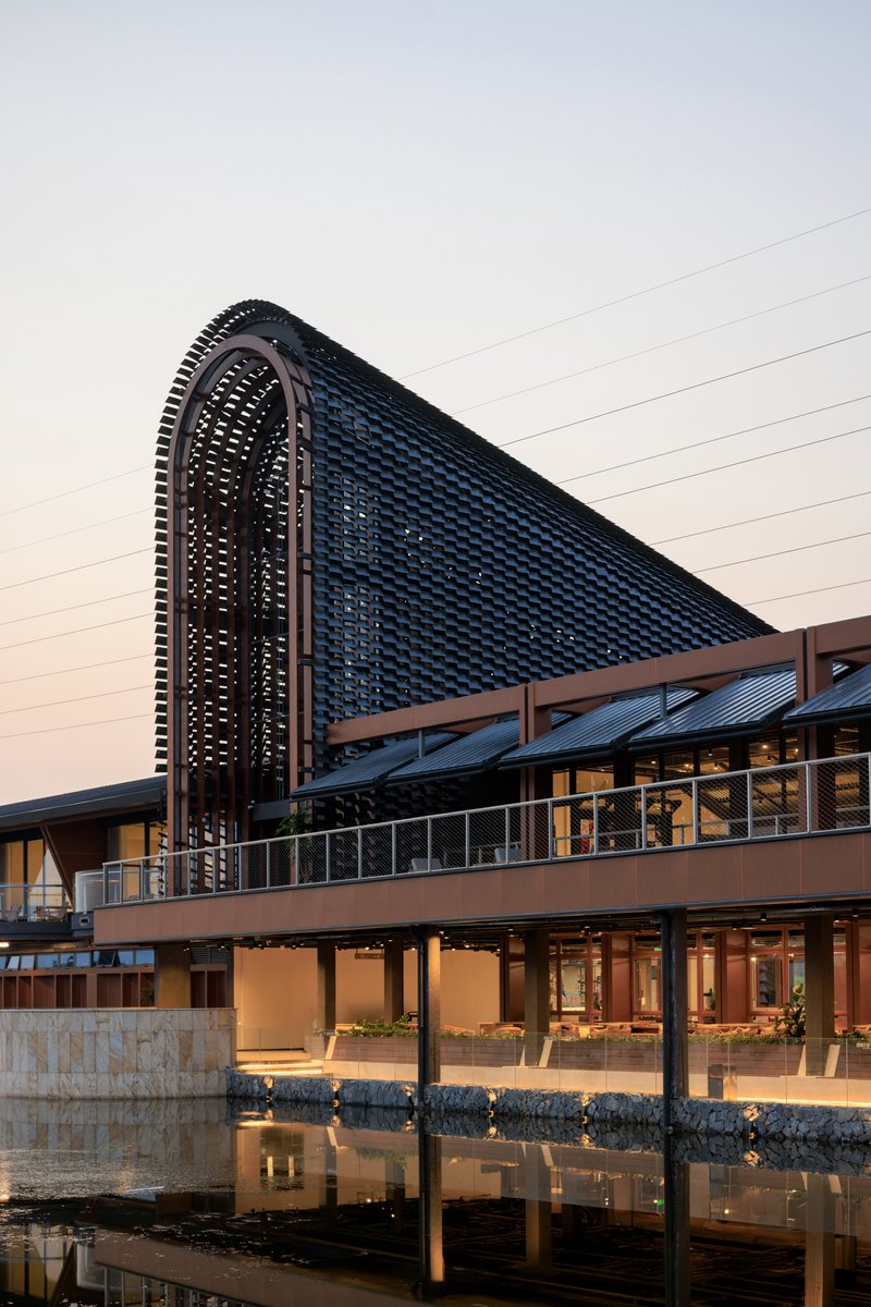 Waterside facade showing the arched perforated screen rising above the timber-framed pavilion at twilight