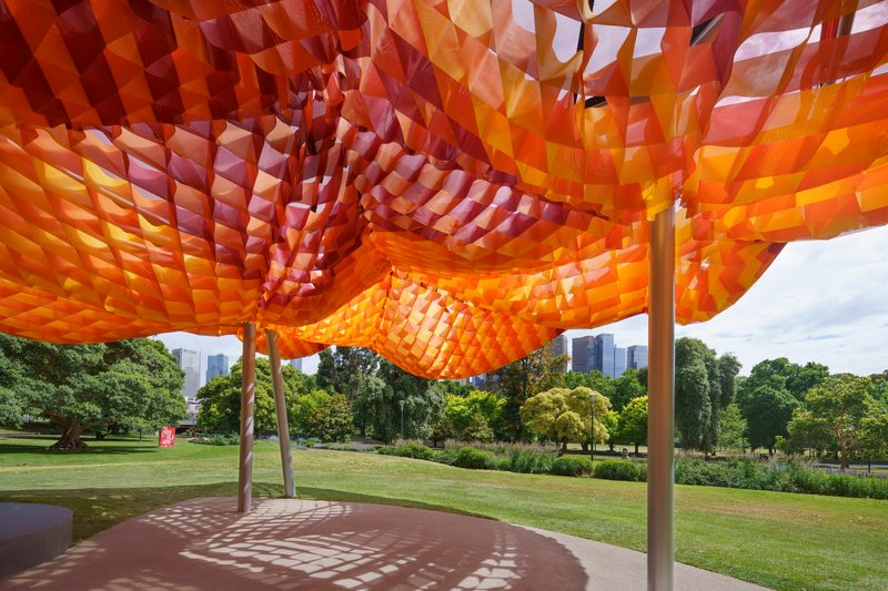 Underside view of the tessellated orange and red fabric canopy with park and skyline beyond