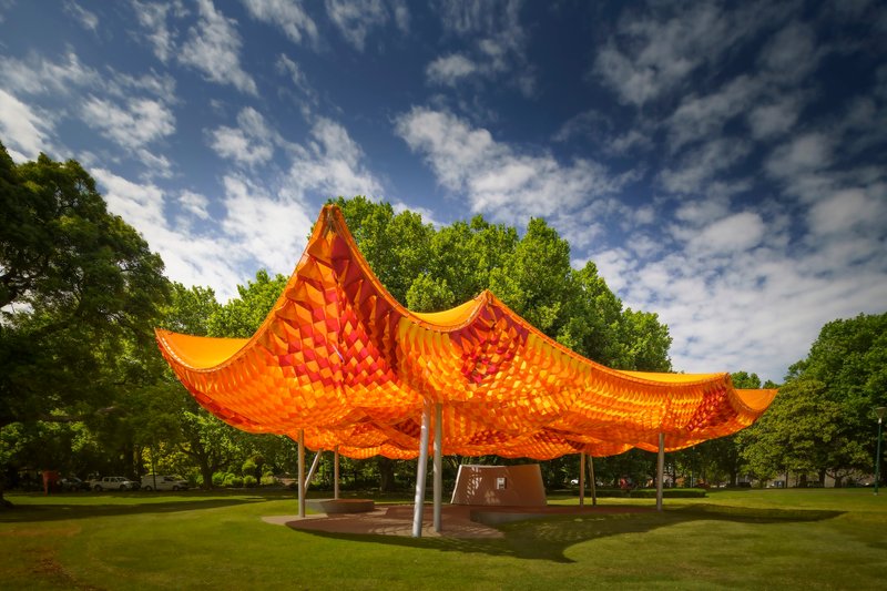 Four-pointed woven canopy pavilion set in a lawn framed by mature trees under blue sky